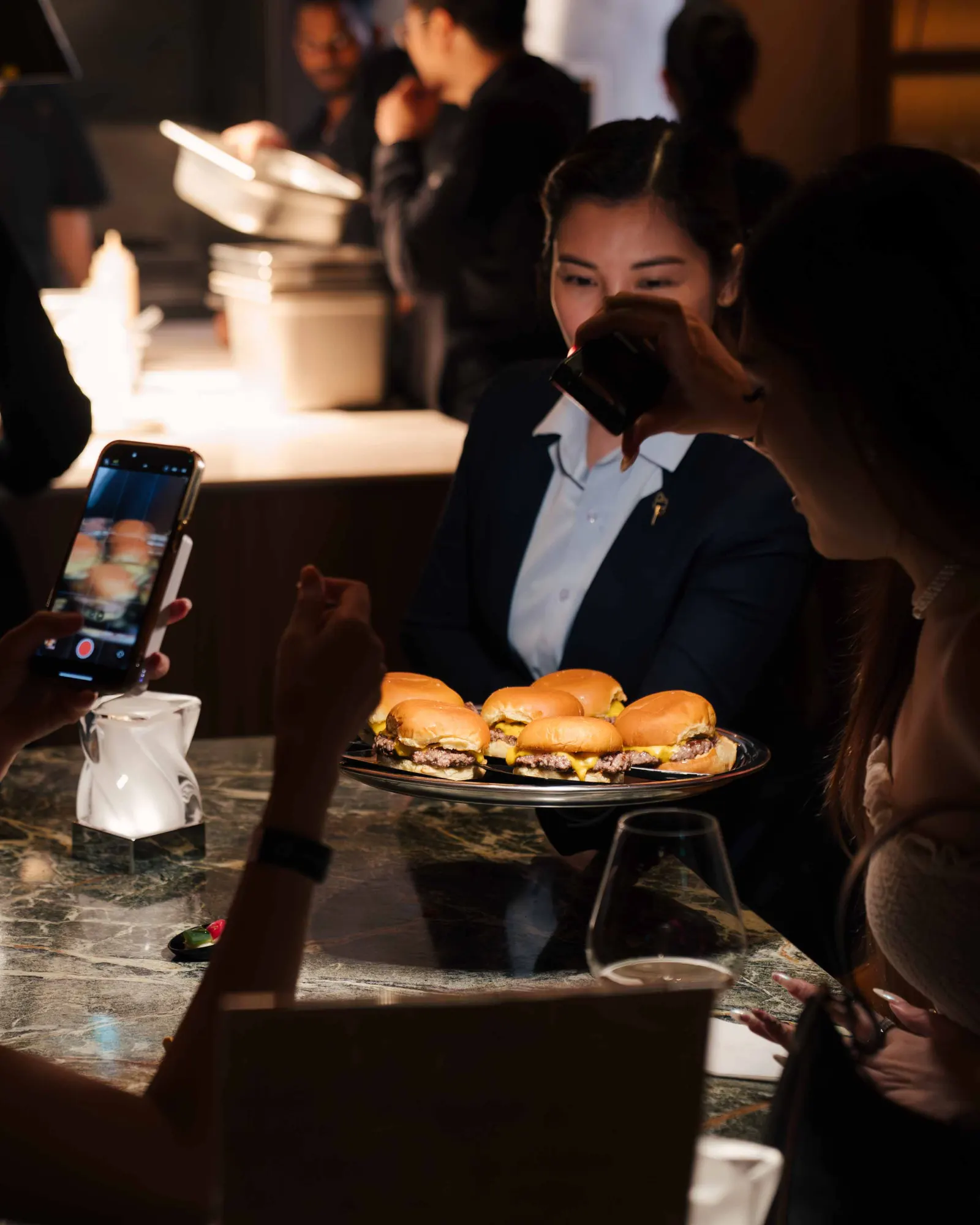 Guest filming a tray of cheeseburger sliders on a phone at the bar while a server looks on