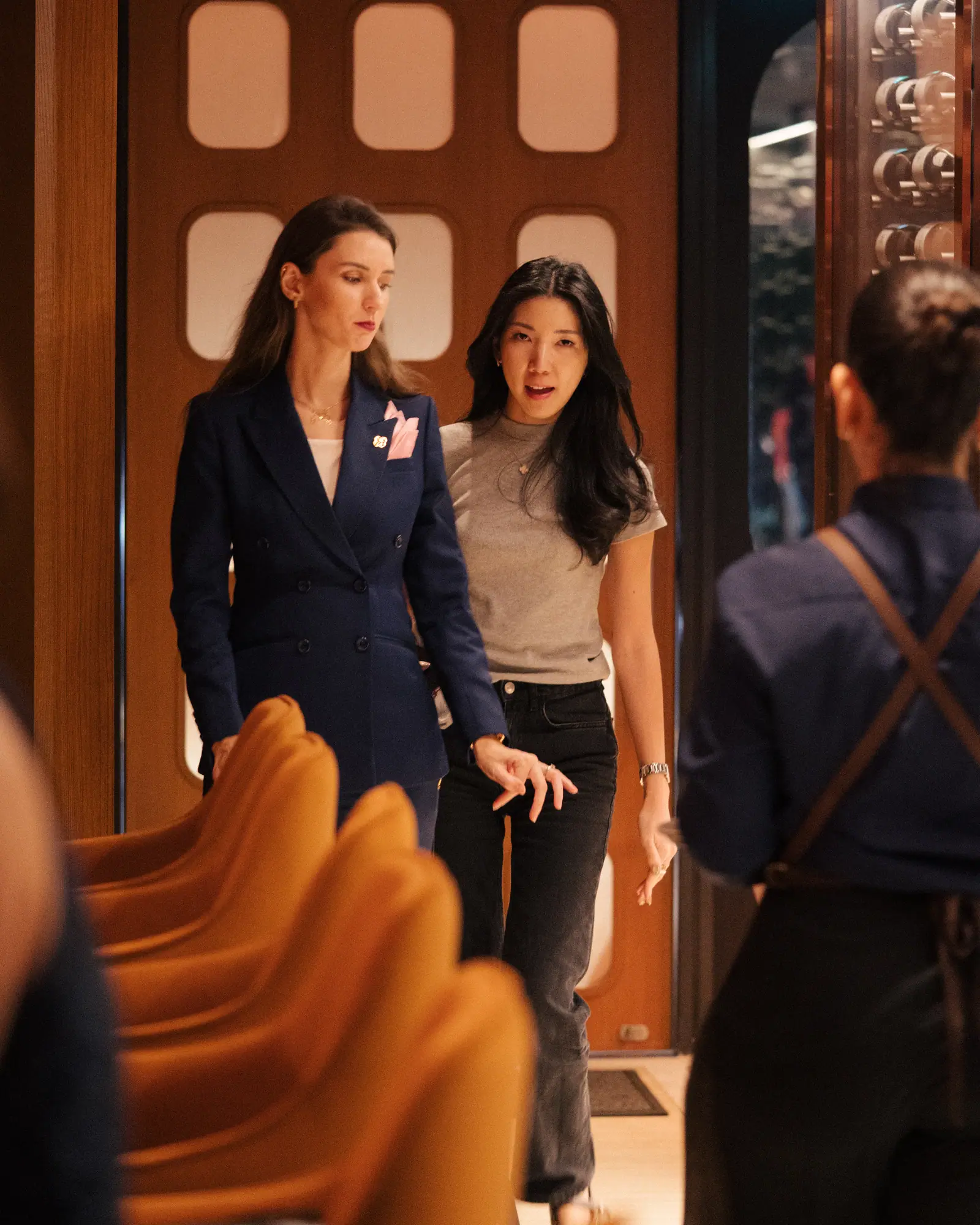 Two women walking into the dining room past tan chairs, staff in the foreground
